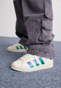 Valkoiset tennarit, joissa on teal- ja purppuraraidat, teksturoitu materiaali ja matala malli. Yhdistetty harmaisiin cargo-housuihin, joissa on taskuja ja ompeleita.