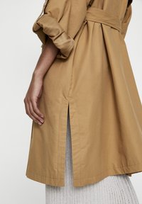 Lichtbruine, lange trenchcoat met een ceintuur in de taille, opgerolde mouwen, zijsplitten en een gladde textuur, gedragen over een geribbeld grijs outfit.