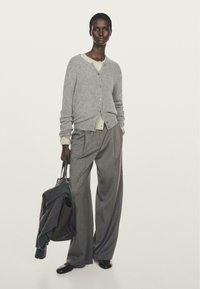 Modella che indossa un cardigan grigio sopra una camicia crema, pantaloni grigi a righe larghe, scarpe nere, mentre tiene una grande borsa a tracolla con un cappotto scuro.