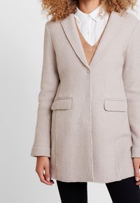 Donna che indossa un cappotto di lana beige chiaro sopra un maglione beige con scollatura a V e una camicia bianca con bottoni, abbinato a pantaloni neri.
