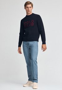 Dunkelblauer Pullover mit rotem "POLO CLUB" Schriftzug, kombiniert mit hellblauen Jeans und beigen Sneakers, für ein legeres Outfit.