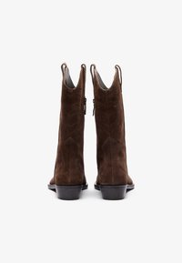 Botas de tobillo de ante marrón con punta afilada, diseño curvado, cremalleras laterales y un tacón apilado con patrones de costura detallados.