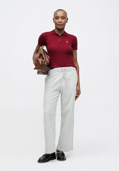 Sieviete, tērpusies bordo īsām piedurknēm polo krekliņā, gaiši pelēkās sporta biksēs, melnās loferēs, tur rokā lielu brūnu ādas rokassomu uz pleca.