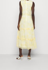 Gul mönstrad maxiklänning med lager på lager - volanger; lättviktsmaterial, ärmlös design och en slät dragkedja i ryggen. Åtföljs av vita klackar.