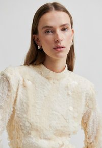 Kobieta z prostymi brązowymi włosami nosi srebrne kolczyki w kształcie koła oraz teksturowany kremowy top z dużymi okrągłymi cekinami, na jednolitym szarym tle.