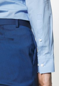 Gros plan d'une personne portant une chemise bleu clair rentrée dans un pantalon de costume bleu foncé avec une ceinture assortie.