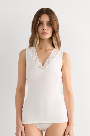 Femme portant un haut blanc sans manches avec une bordure en dentelle sur un col en V et un sous-vêtement blanc assorti, debout devant un fond clair uni.