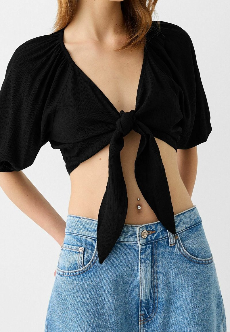 Haut court noir avec lien à nouer sur le devant, en tissu texturé avec manches bouffantes, associé à un short en denim bleu clair taille haute.