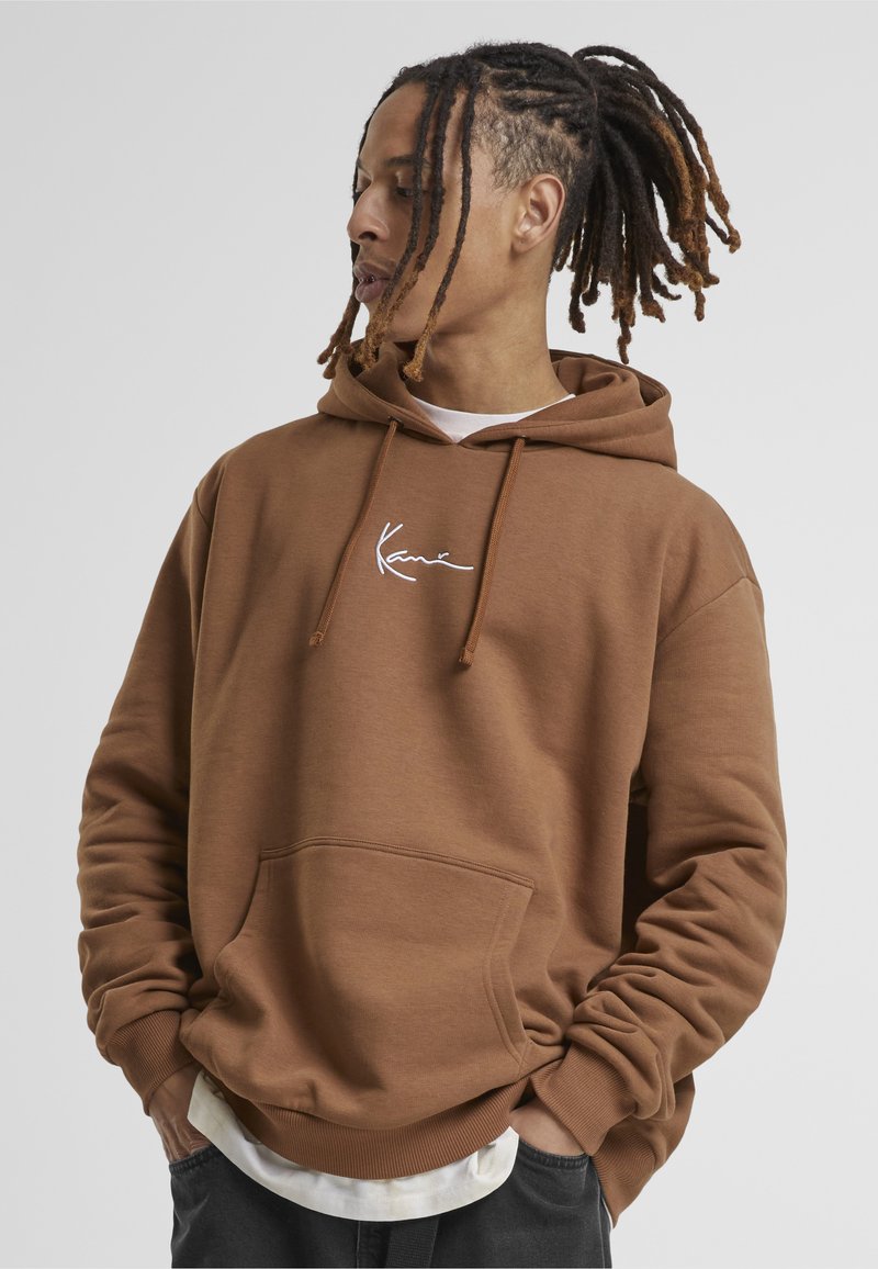 Bărbat cu dreadlocks maro, purtând un hanorac maro cu glugă și logo alb, cu mâinile în buzunare, privind spre stânga pe un fundal simplu.