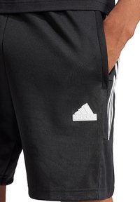 Schwarze Sportshorts aus glattem Stoff. Mit einem weißen Adidas-Logo, Seitentaschen und weißen Streifen an den Seiten.