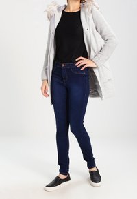 Grau gestrickte Jacke mit Kunstfellbesatz, schwarzes Oberteil, dunkelblaue Skinny-Jeans und schwarze Slipper. Das Model steht vor einem neutralen Hintergrund.