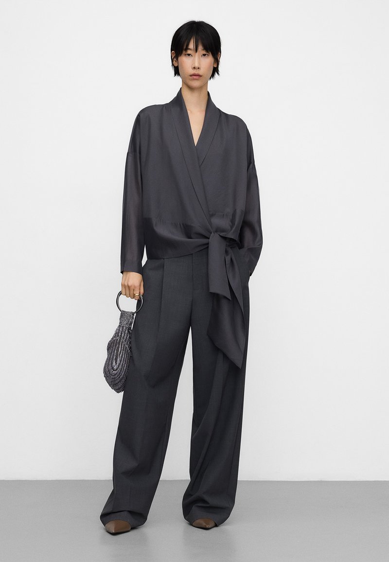 Woman wearing dark gray wrap blouse with side tie, wide-leg trousers, holding small beaded handbag with round metal handle, standing on gray floor.