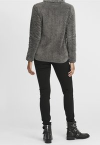 Harmaa tekoturkis takki, jossa on kaulus, yhdistettynä mustiin slim-fit-housuihin ja mustiin soljilla varustettuihin nilkkureihin. Teksturoitu, lämmin ilme.