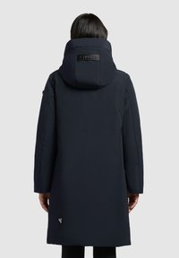 Marineblauwe pufferjas met een grote, gewatteerde capuchon, logo-patch op de achterkant, en een rechte snit met lange mouwen en zijzakken.