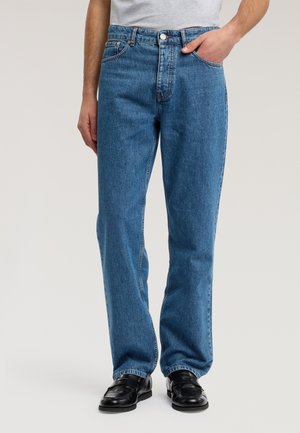 Uomo che indossa jeans di denim blu larghi, mocassini neri in pelle e una maglietta grigia chiara infilata nei pantaloni, con una mano in tasca.