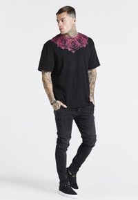 Camiseta negra de manga corta con un estampado de rosa rosa en el pecho, combinada con jeans negros y zapatillas negras. Textura suave, corte relajado.