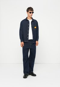 Denim jacket and pants in dark blue, featuring a chest pocket with a yellow label. Model wears sunglasses and black shoes. Smooth texture.