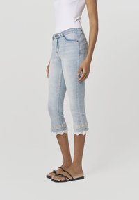 Jasnoniebieskie dżinsowe spodnie capri z ozdobnym koronkowym wykończeniem na dole. Posiadają przednie kieszenie i dopasowany krój, zestawione z czarnymi sandałami.