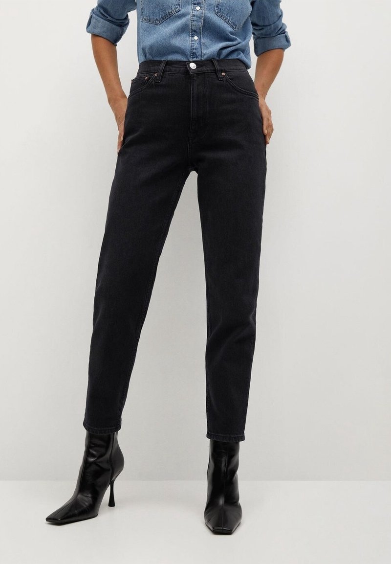 Personne portant une chemise en denim avec manches retroussées, un jean noir taille haute et des bottines à talons noires à bout pointu, debout devant un fond blanc.
