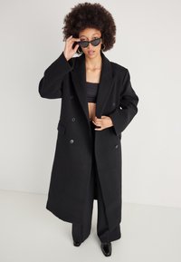 Sort oversized dobbelbrystet frakke med brede ærmer, strukturerede skuldre og knapdetaljer, parret med en sort bralette og brede bukser.
