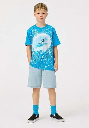 Blauwe grafische t-shirt met spetters en een surferontwerp, gecombineerd met lichte denim shorts, blauwe sokken en zwarte instap schoenen.