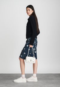 Zwarte geribbelde trui, spijkerrok met bloemenborduurwerk, witte gewatteerde handtas en witte sneakers met textuuroverdes, staand op een grijze vloer.