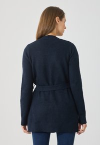 Tmavomodrý pletený cardigan s opaskom v páse, dlhými rukávmi a rebrovaným lemom. Mäkka textúra s minimálnym detailom na chrbte.