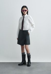 Bijela košulja na dugmad s džepom, tamnosmeđa kravata i crna koža omotačka suknja. Nositi uz srebrne čarape i crne čizme do potkoljenice s detaljima.