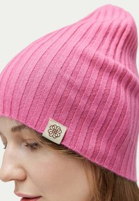 Rosa ribbatten stickad mössa med en passande form. Har en liten krämfärgad etikett med en brun dekorativ design på sidan. Mjuk textur.