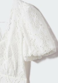 Blouse blanche à manches courtes bouffantes, avec une broderie en dentelle complexe et des détails en œillets, créant un effet texturé et respirant.