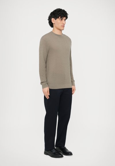 Svetlohnedý sveter s textúrou pleteniny s okrúhlym výstrihom, rebrovanými manžetami a lemom, v kombinácii s tmavomodrými nohavicami a čiernymi topánkami.