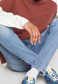 Brun kortärmad tröja över en krämfärgad långärmad tröja, kombinerad med ljusblå trasiga jeans och flerfärgade mönstrade sneakers.