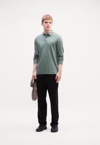 Long-sleeve, light green polo shirt with a collared neck, paired with black trousers and black sneakers. Model holds a gray shoulder bag.