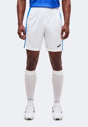 Athlète masculin portant un short de sport blanc avec des rayures bleues, des chaussettes blanches hautes et des crampons, debout les mains détendues le long du corps.