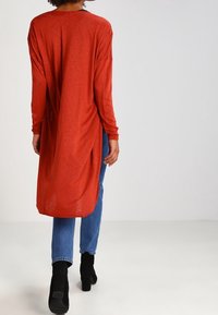 Langärmliges, locker sitzendes Oberteil in einer strukturierten Rostfarbe, mit einem High-Low-Saum, kombiniert mit blauen Jeans und schwarzen Stiefeletten.