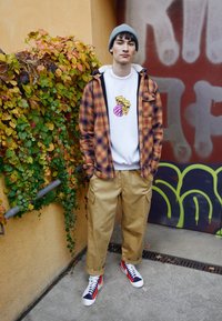 Kostkovaná flanelová bunda přes šedou mikinu s grafikou, tanové cargo kalhoty a barevné tenisky; zeleň v pozadí.