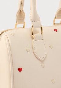 Τσάντα σε κρεμ χρώμα από συνθετικό δέρμα με χρυσά μεταλλικά στοιχεία, διακοσμημένη με μικρές διαμορφωμένες καρδιές σε κόκκινο και κρεμ χρώμα στην επιφάνεια.