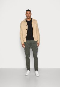 Helles beiges Leinenhemd, schwarzes T-Shirt, olivfarbene Cargo-Hosen mit Taschen, weiße Sneakers, stehend vor einem schlichten weißen Hintergrund.