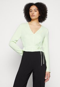 Top verde claro de mangas longas com decote em V e detalhe de cordão na cintura, emparelhado com calças largas pretas. Textura suave.