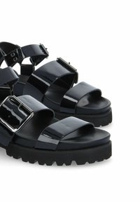 Sandales en cuir verni noir avec une semelle en caoutchouc épaisse, de larges sangles croisées et un grand accent en boucle argentée sur la sangle de la cheville.