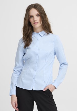 Ung kvinde med langt brunt hår iført en lyseblå skjorte med knapper og sorte bukser, stående med den ene hånd i lommen.