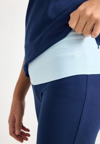 Dunkelblaues Sportoberteil über einer hellblauen Basisschicht getragen, kombiniert mit dunkelblauen Leggings. Der Stoff wirkt glatt und dehnbar.