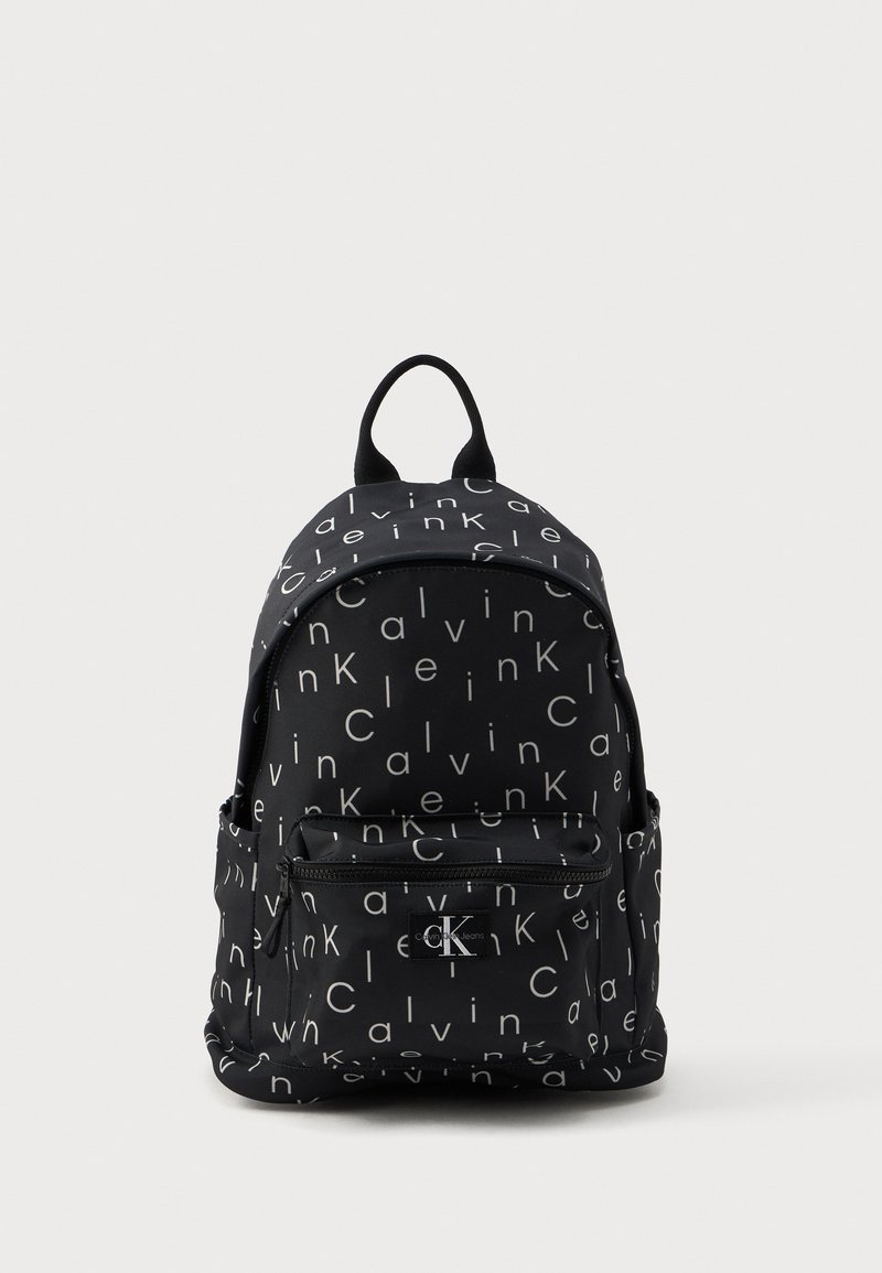 Μαύρο σακίδιο διαθέτει επαναλαμβανόμενο σχέδιο κειμένου με την ένδειξη "Calvin Klein" σε λευκό. Περιλαμβάνει μπροστινή τσέπη με φερμουάρ και μία λαβή στην κορυφή για μεταφορά.