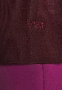 Bordeaux geribbeld breiwerk met een geborduurd "VVB" logo, gecombineerd met een gladde, levendige paarse stof daaronder, wat zorgt voor een contrasterende textuur.