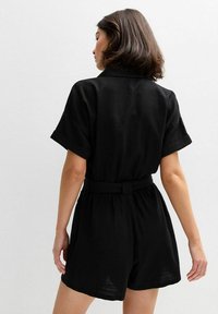 Svart kortärmad jumpsuit med bälte i midjan, med en krage och avslappnad passform, tillverkad av lätt texturerad tyg.
