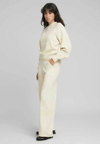 Sudadera color crema con bolsillo frontal y corte holgado, combinada con pantalones de pierna ancha a juego. Textura suave, diseño minimalista, puños acanalados.