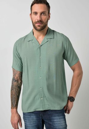 Mann trägt ein kurzärmeliges, hellgrünes, gemustertes Hemd mit Knöpfen, blaue Jeans, Armbanduhr und sichtbares Arm-Tattoo, steht vor einfärbigem Hintergrund.