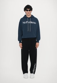 Námořnická mikina s nápisem "No Problemo", šňůrkou, a černé tepláky s potiskem "No Problemo". Bílé tenisky, hladká textura.