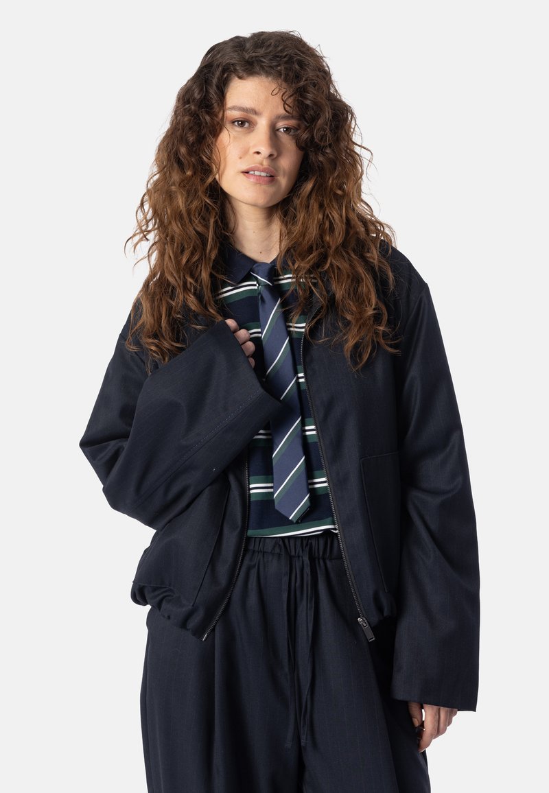 Femme aux longs cheveux bruns bouclés portant une veste sombre surdimensionnée, une chemise rayée et une cravate, debout devant un fond clair uni.
