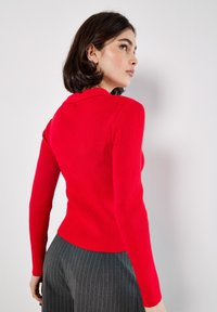 Czerwona sweter z długim rękawem, prążkowany, z kołnierzykiem, dopasowana sylwetka i teksturowana tkanina, zestawiony z szarymi spodniami w prążki.